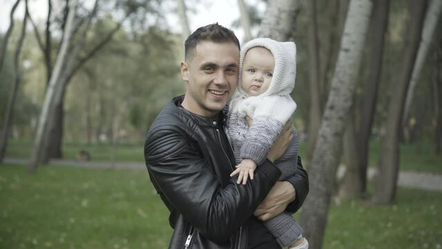 Happy handsome young man kissing infant and looking at camera smiling. Portrait of cheerful Caucasian father enjoying day outdoors with cute charming son holding on hands.