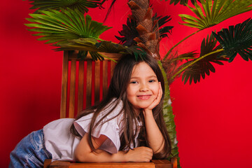 A little girl stands next to a palm tree on a solid red background. Portrait of a child with a beautiful smile.