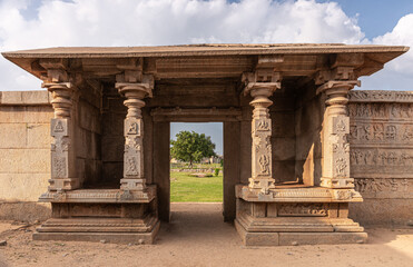 Fototapeta premium Hampi, Karnataka, India - November 4, 2013: Hazara Rama Temple. Beige stone entrance hall with flat roof and fresco decorated pillars is set in outside wall, showing green foliage and blue cloudscape.