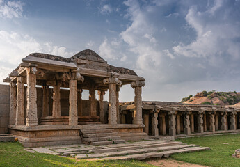 Hampi, Karnataka, India - November 4, 2013: Hazara Rama Temple. Ruinous brown stone buildings along outside walls with rows of columns under blue cloudscape with grass in front.
