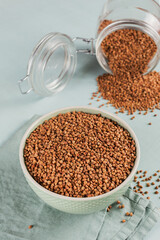 Organic uncooked scattered buckwheat grain in a bowl and glass jar on a light blue background. Healthy and diet food concept.