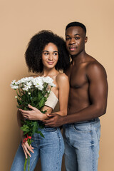 shirtless african american man hugging cheerful woman with flowers on beige