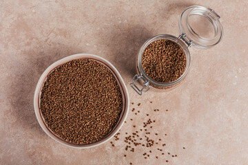 Organic uncooked scattered buckwheat grain in a bowl and glass jar on a light beige background. Healthy and diet food concept.