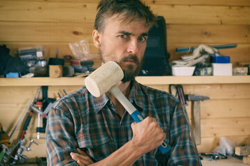 Brutal bearded worker man stands in workshop with mallet, construction carpentry tools in the background.  DIY concept.
