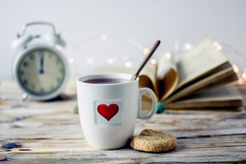 Valentine's day breakfast. White mug of tea with red heart, alarm clock, biscuit heart, garland book on a wooden table. Romantic love background.