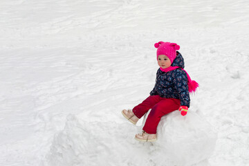 Fototapeta na wymiar Little caucasian girl in a pink hat sits on a snow ball, winter fun making a snowman