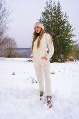 Fototapeta na wymiar Winter lifestyle, portrait of young pretty blond Caucasian woman with white winter outfit and woolen hat in snow next to snowy pine trees, vacation in nature