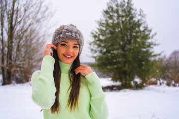 Winter lifestyle, portrait young brunette caucasian with green outfit and woolen hat, smiling in the snow next to snowy pine trees, vacation in nature