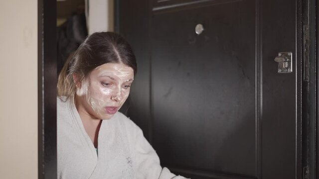 A young woman in a dressing gown opens the front door with a cosmetic mask on her face.