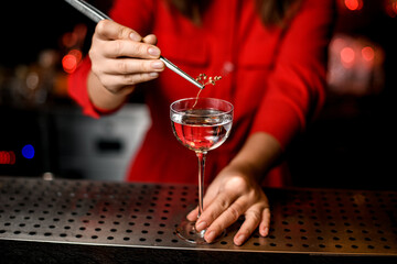 hand of woman holding tweezers with sprig of plant over wine glass on bar counter