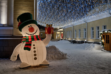 Big colorful funny inflatable decorative snowman on a winter evening illuminated city street.