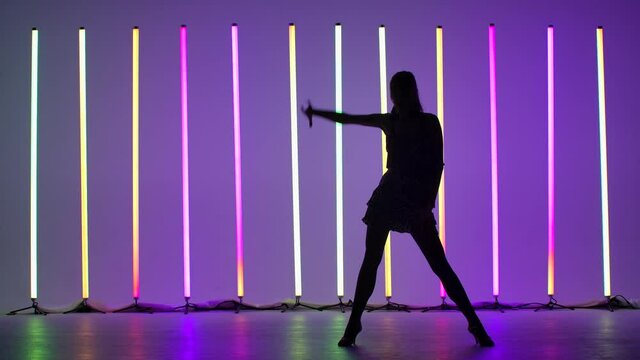 Professional slim dancer dancing rumba in the studio against the backdrop of bright multicolored neon lights. The girl works out the steps of the dance. Silhouette. Slow motion.