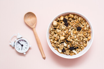 Healthy eating. Baked oats, nuts and raisins granola in a bowl, alarm clock and a wooden spoon on a pink background. Top view