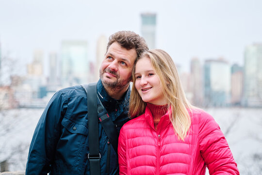 A pretty teenager and her father are posing for the camera. They are wearing puffy coats and look cosy and warm. They look happy, their lips are smiling. The New York Skyline is in the background.