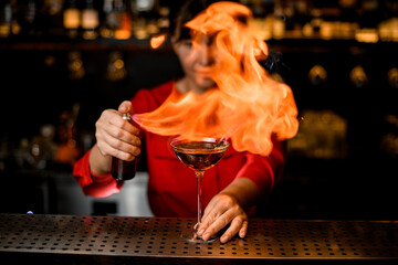 wine glass with drink stands on bar and lady barman masterfully sprinkles and sets fire flame over it