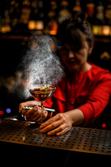 wine glass with brown drink stands on bar counter and female bartender sprinkles over it
