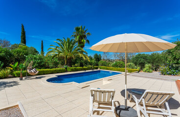 Pool with poolside umbrella and sun loungers. Clear blue sky and beautiful trees in the background on a nice summer day.