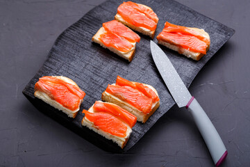 Bruschettes with butter and trout on a black wooden board on a gray background near a knife.
