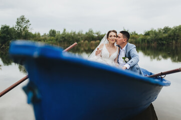 Stylish groom in a suit and a cute bride in a white lace dress are sitting in a wooden boat, walking and swimming on the lake, enjoying the beautiful nature.