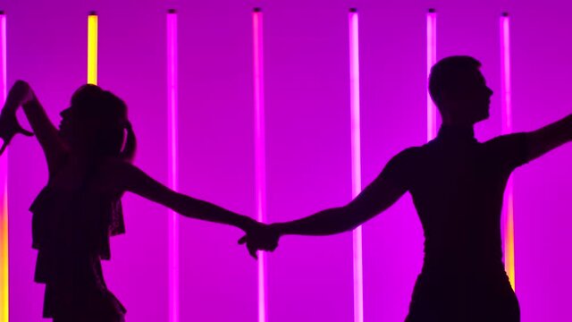 A couple of young dancers are dancing a passionate Latin American dance. Rumba, samba, cha cha cha, jive. Dark silhouettes of two dancers against the background of neon lamps. Slow motion. Close up.