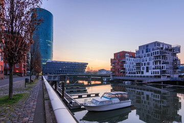 Cityscape in Frankfurt am Main with canal and buildings. Boats in the harbor before sunrise in the West Harbor residential area. Road with trees and reflection in the water