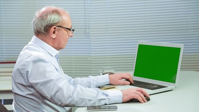 A man with glasses in an office with blinds at a computer prints a green screen and writes in a notebook. Medium plan
