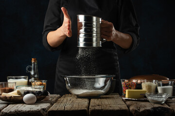 Pastry chef sifts flour into glass bowl on wooden table with variety of ingredients on dark blue background. Backstage of preparing sweet waffles. Delicious dessert. Frozen motion. Cooking process.