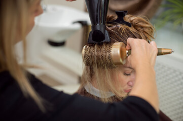 Closeup of hairdresser brushing woman's blond hair using a hair dryer and professional round brush