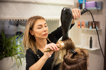 Fototapeta premium Elegant mature hairdresser enjoying working in beauty salon and blowdrying hair of client