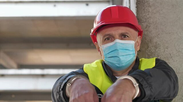Handcuffed worker in red hard hat and medical protective mask at construction site, he looks at the camera