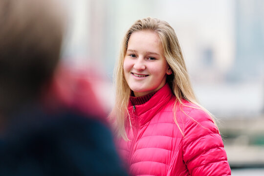 A pretty teenager is wearing a pink puffy coat and looks cosy and warm. She looks happy, has long fair hair and white skin with rosy cheeks and green eyes. The New York Skyline is in the background.