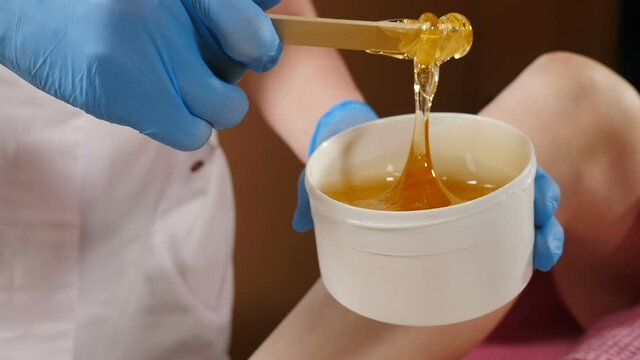 Beauty concept. Sugar paste in white jar. Beautician in blue gloves stirs sugar paste before removing hair. Sugaring. Depilation. Epilation. Paste flows down from wooden waxing spatula sticks. Full hd