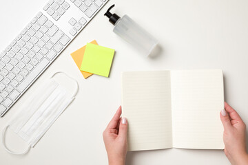 Top view of a white table with a keyboard, a white surgical mask, two colored reminder stickers, a alcohol gel pump dispenser and female hands holding a blank notebook. With copy space.