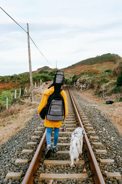 unrecognizable musician in a yellow coat walking her dog along the train tracks with the guitar loaded on his back. youth concept