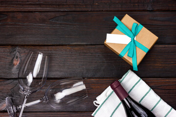 Gift wrapped in craft paper, a bottle of red wine and two glasses on a dark wooden table. View from above. A gift for a loved one.