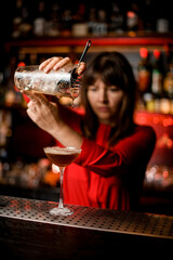 wineglass with cocktail stands on bar and woman bartender holds mixing cup over it