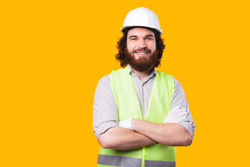 A awesome portrait of a young bearded engineer looking at the camera with his arms crossed near a yellow wall .
