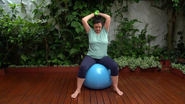 Mujer haciendo ejercicio con mancuernas sentada en una pelota  en el jard&iacute;n de su casa