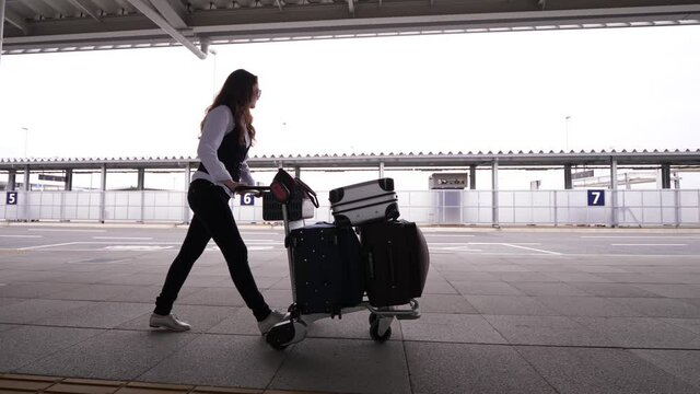 Woman push baggage dolly full of suitcases, walk near airport terminal. European lady arrive to or depart from domestic terminal of Japanese airport. Tracking shot, side view