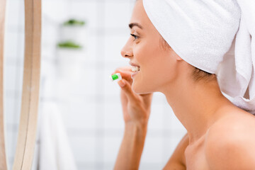 side view of woman with white towel on head brushing teeth in bathroom near mirror