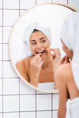 young woman with white towel on head flossing teeth near mirror in bathroom, blurred foreground