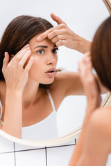 Fototapeta premium displeased young woman touching face while looking in mirror, blurred foreground