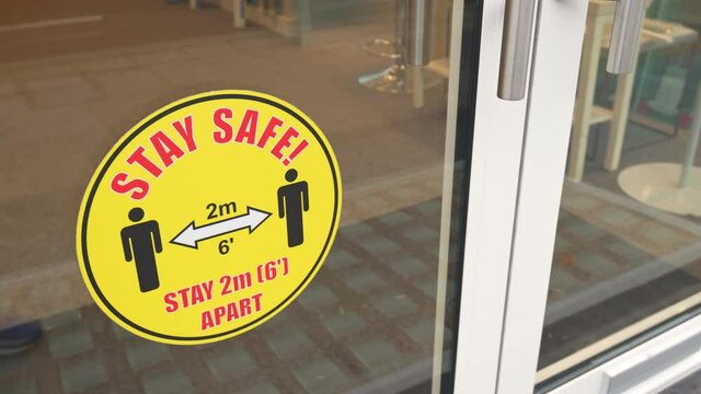 Stay Safe 2m 6′ apart: warning sticker banner on the door at the store entrance. Guidance for people during Coronavirus COVID-19 pandemic in 2021.