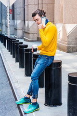 Young Hispanic American with hair bun, wearing glasses, yellow long sleeve T shirt, blue jeans, green sneakers, holding cup of coffee, sitting on street in New York City,  talking on cell phone..