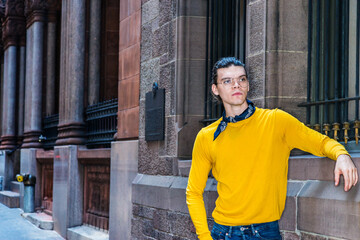 Young Hispanic American Man with hair backward, wearing glasses, yellow long sleeve T shirt, small black scarf around neck, standing by old style wall with windows on street in New York City..