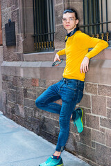 Young Hispanic American Man with hair backward, wearing glasses, yellow long sleeve T shirt, blue jeans, green sneakers, small black scarf around neck, standing by old wall on street in New York City.
