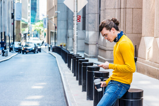 Young Hispanic American with hair bun, wearing glasses, yellow long sleeve T shirt, blue jeans, small black scarf around neck, holding cup of coffee, sitting on street in New York City, texting..