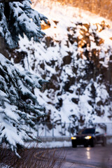 Snowy fir trees on focus, with a black vehicle, snowy rocks and forest lit by the sun in soft focus in the background