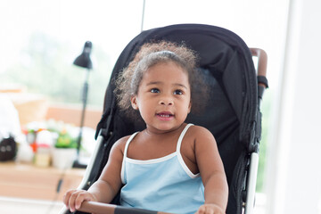 African American little girl with curly hair sitting and playing on stroller. Cheerful Afro toddler little girl sitting on stroller. Toddler child little girl enjoy, having fun at home