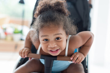 African American little girl with curly hair sitting and playing on stroller. Cheerful Afro toddler little girl sitting on stroller. Toddler child little girl enjoy, having fun at home
